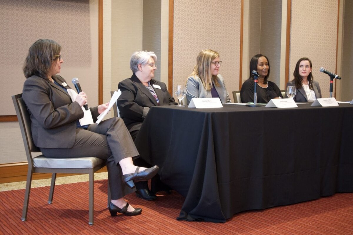 4 women sitting at a table for a panel discussion, 1 woman sitting separately to interview the panel. 